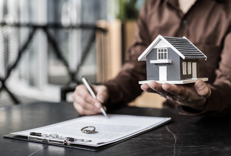 A person holding a small house model in one hand while signing a document on a clipboard with the other hand, symbolising a real estate transaction or property agreement.