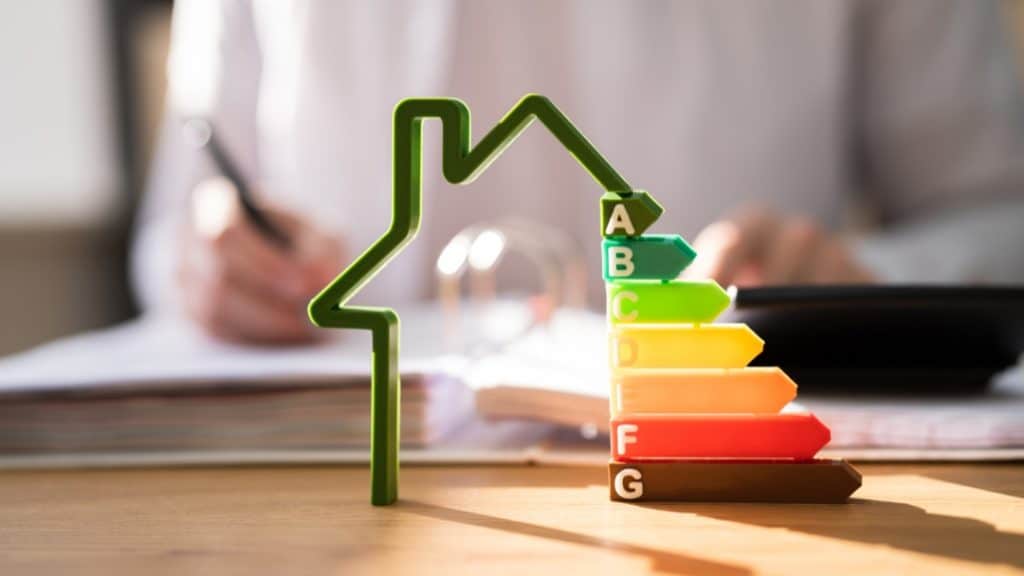 A person working at a desk with a pen and calculator in the background, and a model of a house with a colourful energy efficiency rating chart ranging from A to G in the foreground.