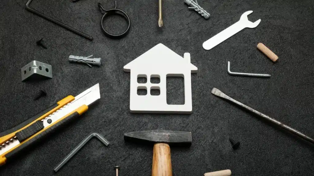 A white house-shaped cutout surrounded by various tools and hardware items, such as a hammer, utility knife, screws, and wrenches, symbolising home improvement or construction projects.
